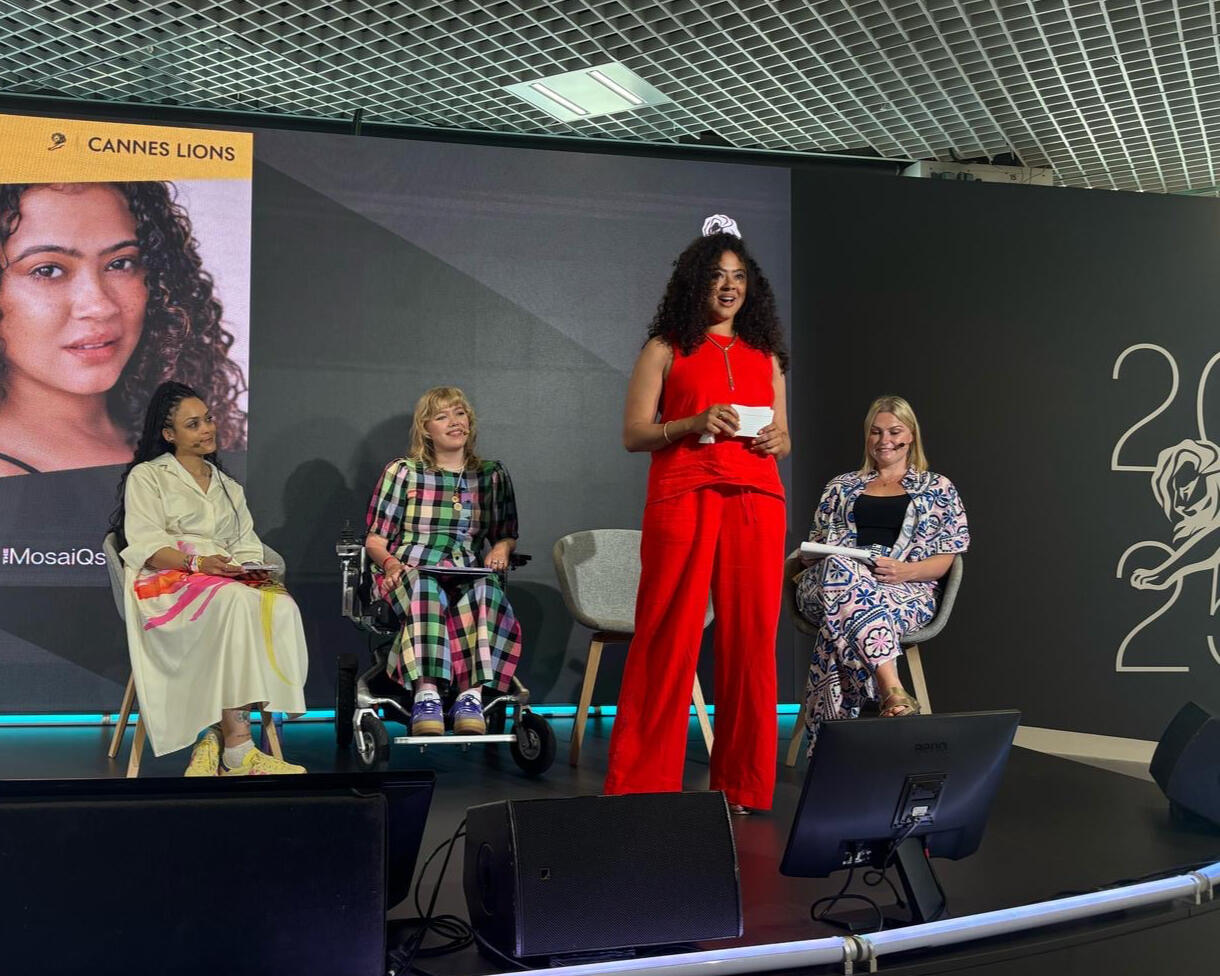 Natalie Amber wearing a Red co-ord, trouser set, standing on the Forum stage, in front of Charlotte, Emma and Laura who are seated behind her, she is addressing the audience.