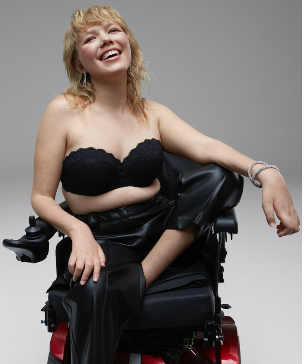 Emma Lines Portrait of Emma Lines, a model and disability advocate, seated in a power wheelchair. She wears a black bandeau top and trousers, smiling brightly. Her bio highlights her use of fashion to challenge norms and push for visibility in the industry.